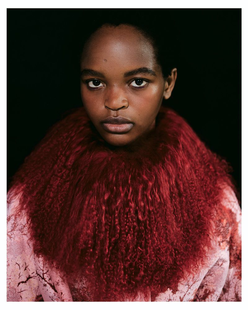 Close-up portrait of a model with a serious expression wearing a crimson fur collar against a solid black backdrop.