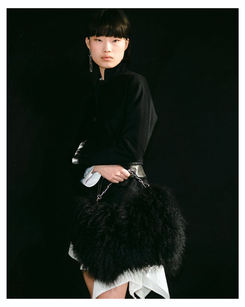 East Asian model holding a large black fur handbag wearing a structured black jacket against a solid black seamless background.