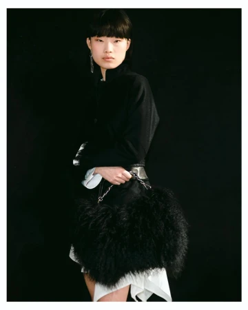 East Asian model holding a large black fur handbag wearing a structured black jacket against a solid black seamless background.