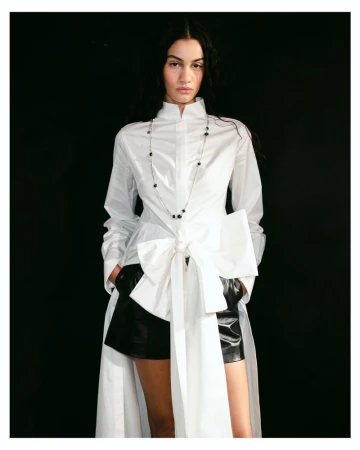 Model wearing a white high-neck tunic with an oversized front bow and black shorts against a solid black backdrop.