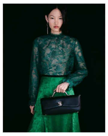 Model wearing a sheer green lace top and matching skirt holding a black leather handbag against a black background.