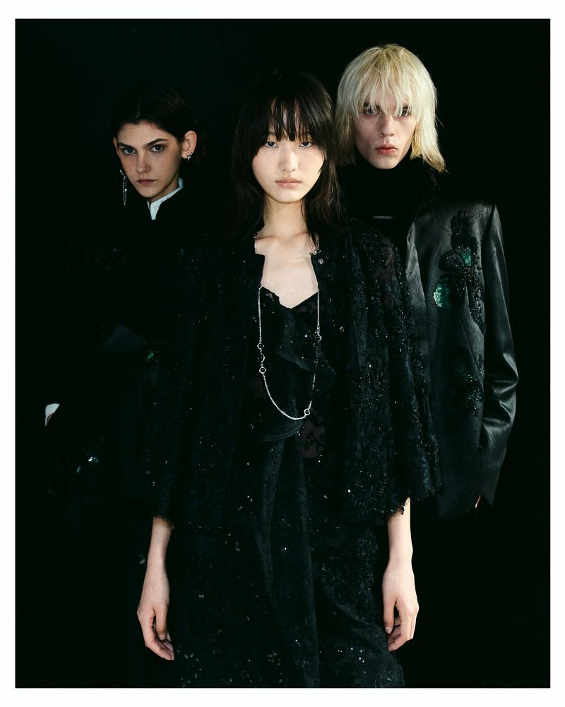 Three fashion models wearing dark textured garments and jackets posing against a solid black background.
