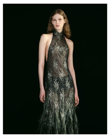 Model posing in an intricately embroidered sheer halterneck evening gown with feathered hem detailing against a black backdrop.