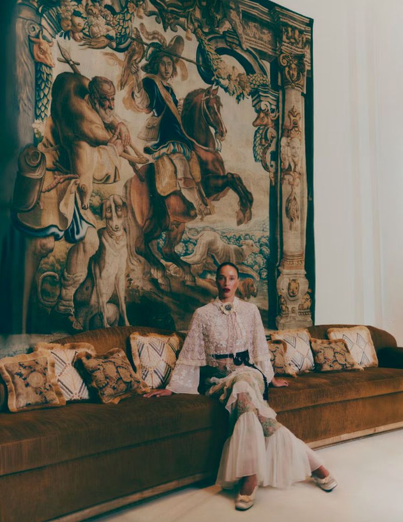 Fashion editorial shot of a woman in a white lacy dress sitting on a brown couch in front of a large tapestry indoors.