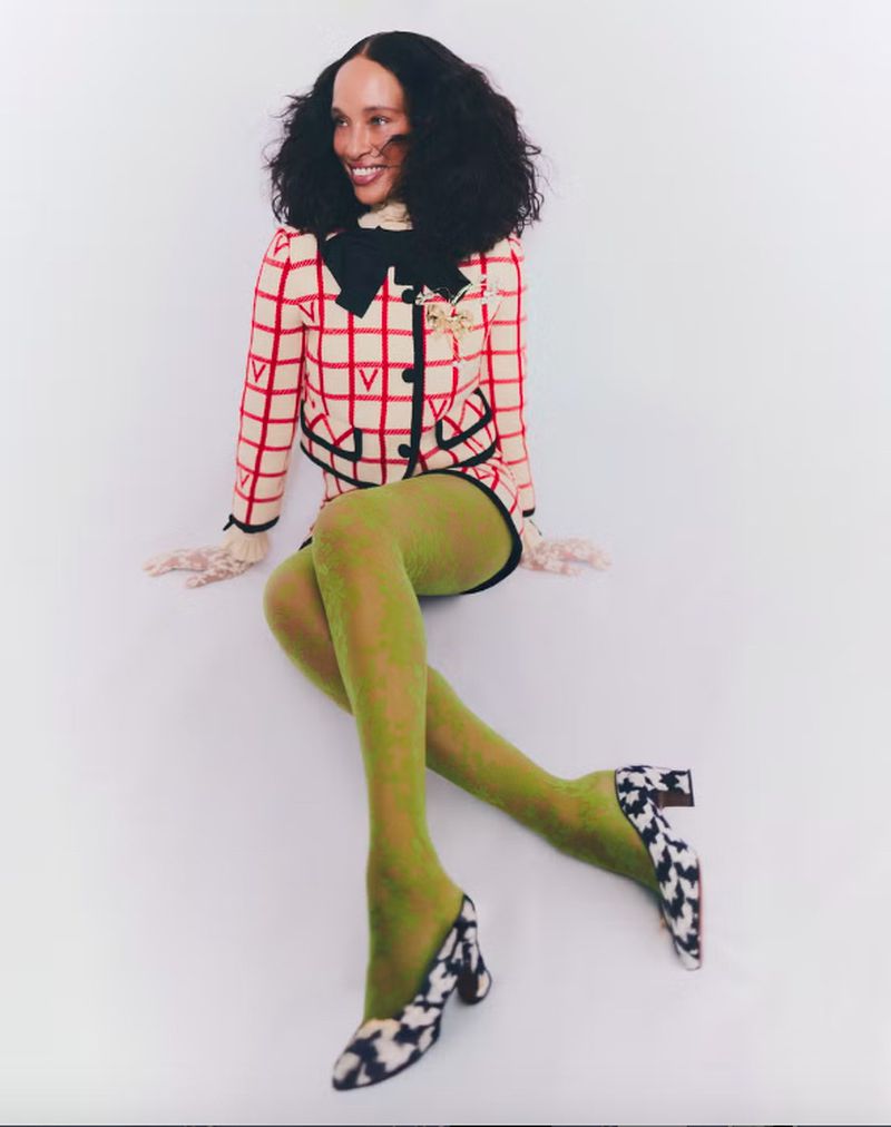 Smiling woman in studio wearing checked jacket, green tights and printed pumps sitting against white background in fashion portrait.