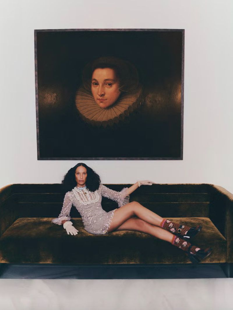An adult woman is reclining on an olive velvet sofa in front of a portrait hanging on a neutral wall in an indoor setting.