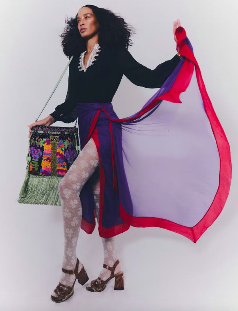 A fashion shot of a woman in a black top, a red and blue wrap skirt, patterned hosiery, and chunky sandals, posing in a studio.