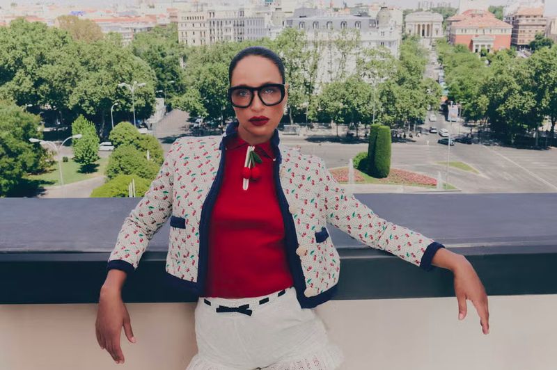 Woman in cherry-print jacket poses on rooftop overlooking city. She wears glasses, shorts, red top, and white jacket with cherry pattern.
