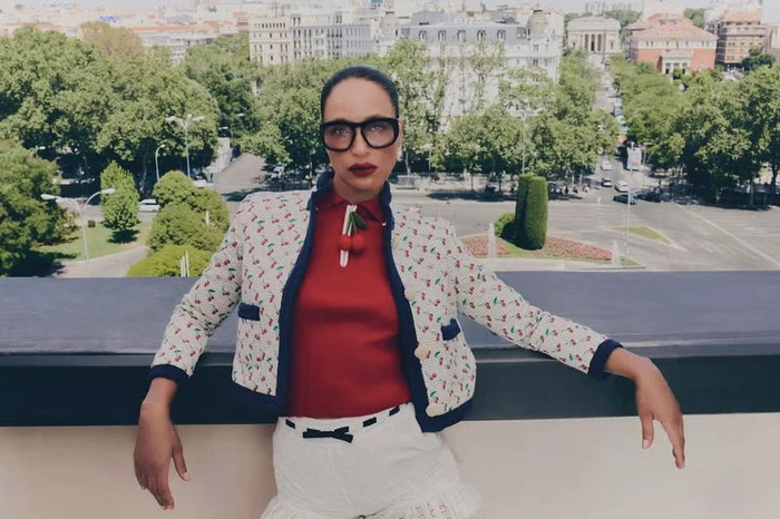 Brandi Quinones wearing thick black glasses, a red top, and a cherry-patterned jacket posing on a rooftop overlooking a city.