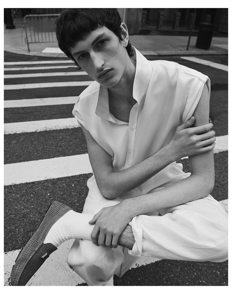 Black and white studio shot of a male model posing in white clothing on a crosswalk, seated with his arms folded, looking at the camera.