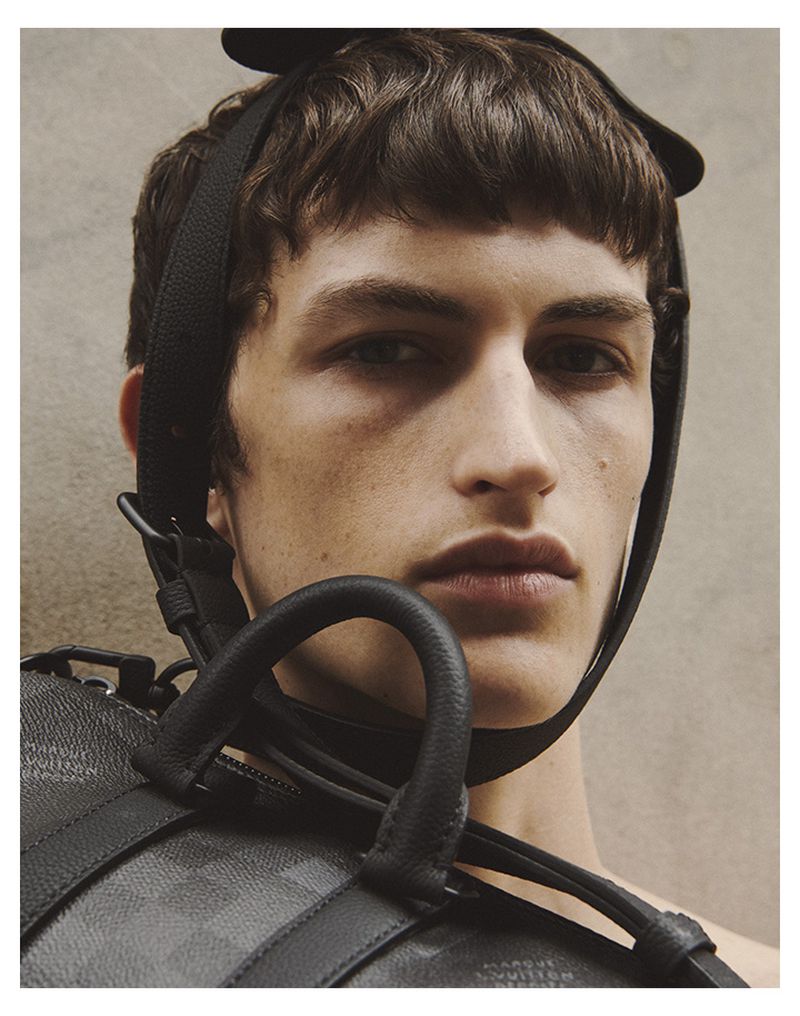Close-up studio shot of a young male model wearing a patterned black leather bag with shoulder strap. Fashion advertising.