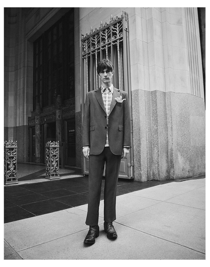Full-length shot of a young adult man in a suit standing in front of a granite building with ornate ironwork details and a city sidewalk.