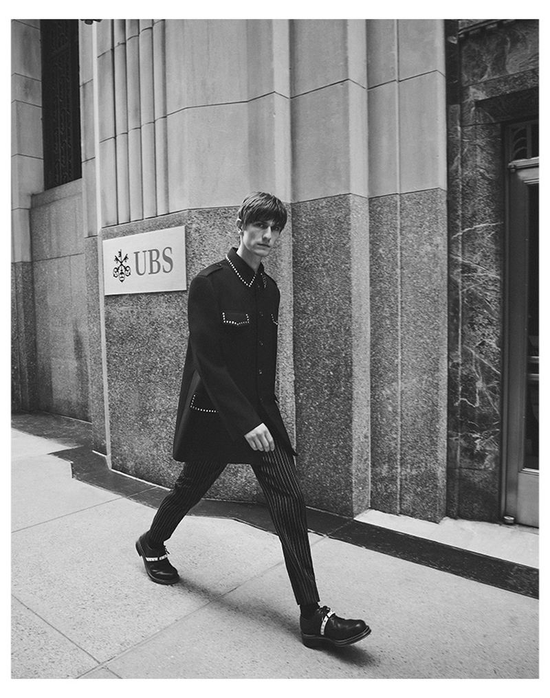 Black and white outdoor fashion shot of a slender adult male walking past a corporate building in a dark jacket and pinstripe pants.
