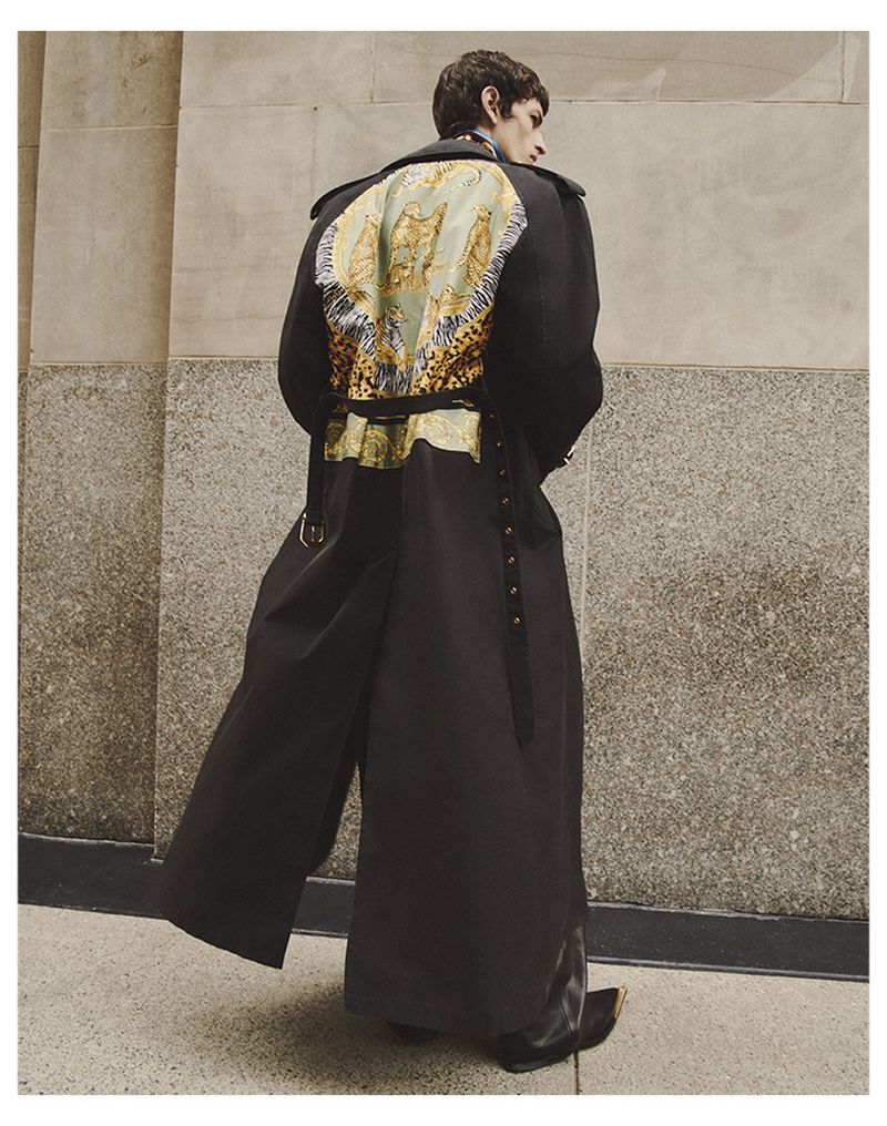Man stands in a black overcoat with an ornate gold and leopard print panel, back to the camera, on a sidewalk in front of a stone wall.