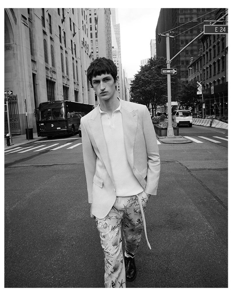 Black and white shot of a male model in a blazer and patterned pants walking on a city street with buildings and traffic.