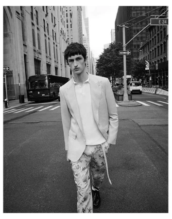 Black and white shot of a male model in a blazer and patterned pants walking on a city street with buildings and traffic.