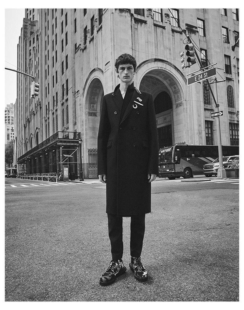 Monochrome fashion shot of a man in a dark coat and patterned shoes standing on a city street with buildings and a traffic light.