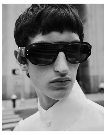 Close-up black and white portrait of a male model wearing large black sunglasses and a high-collared white shirt outdoors.