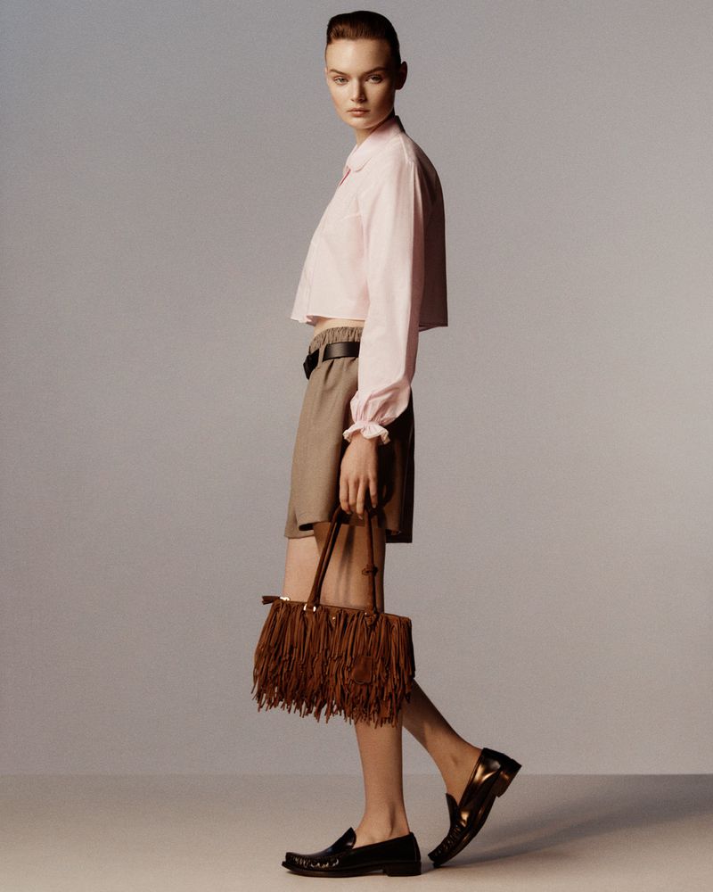 Fashionable young woman in pink blouse, khaki shorts, and black loafers carries a fringed brown leather handbag, studio shot.