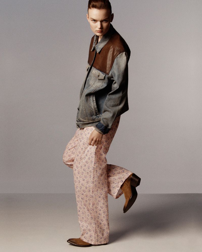 Model Lilli Cummings poses in a denim and leather jacket, pink floral trousers, and brown boots against a grey studio backdrop.