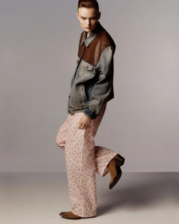 Model Lilli Cummings poses in a denim and leather jacket, pink floral trousers, and brown boots against a grey studio backdrop.