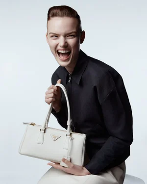 Model Lilli Cummings laughing while holding a structured white Prada handbag in a dark zip-up jacket against a white studio background.