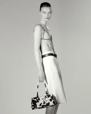 Model Lilli Cummings poses in a triangle bra and white skirt holding a cow-print handbag against a neutral grey studio background.