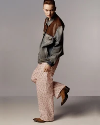 Model Lilli Cummings poses in a denim and leather jacket, pink floral trousers, and brown boots against a grey studio backdrop.