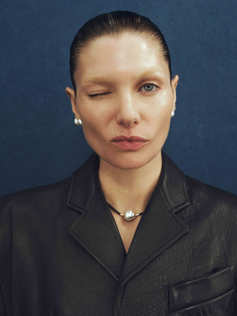 Close-up studio portrait of a fashionable adult woman winking at the camera, wearing pearl jewelry and a black leather jacket.