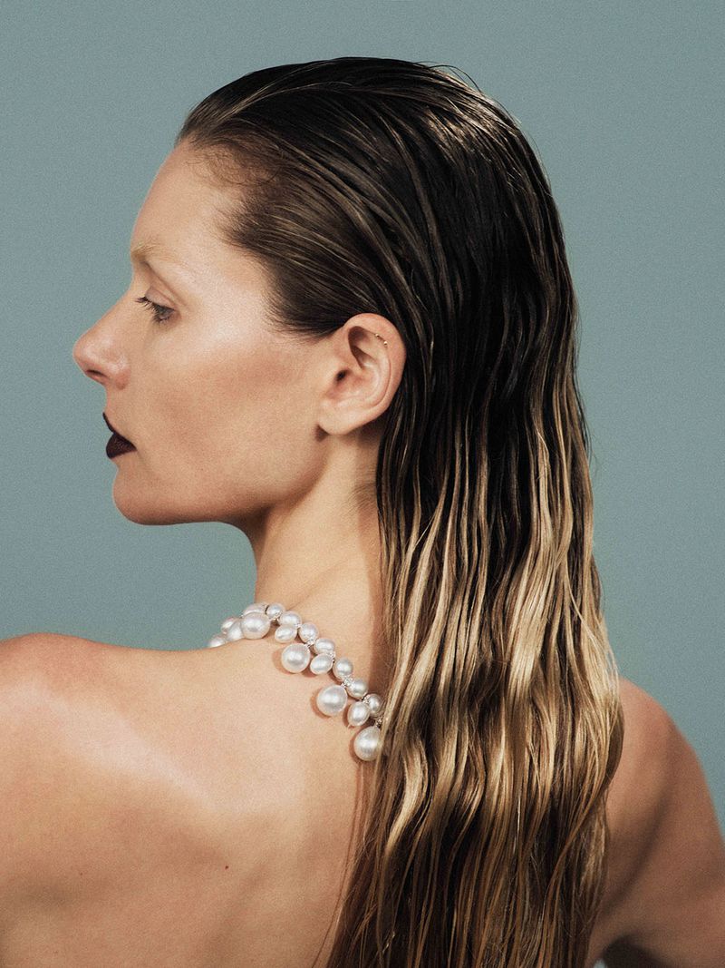 Profile of a woman with wet hair and a pearl necklace against a light blue backdrop, captured in a beauty and fashion-focused studio shot.