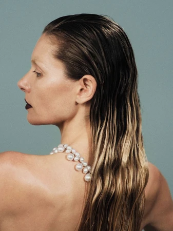 Profile of a woman with wet hair and a pearl necklace against a light blue backdrop, captured in a beauty and fashion-focused studio shot.