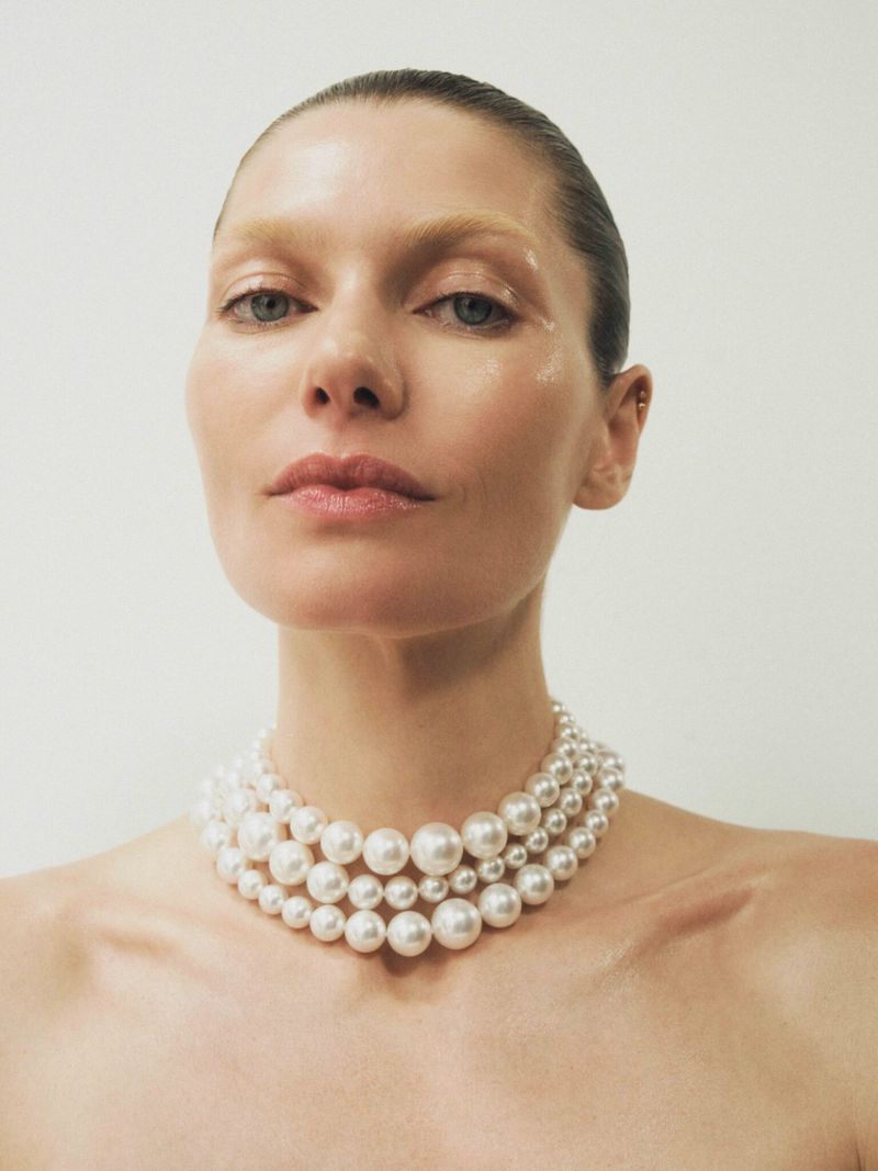Close-up portrait of a woman with light skin, wearing a pearl necklace, and posing in front of a white background.