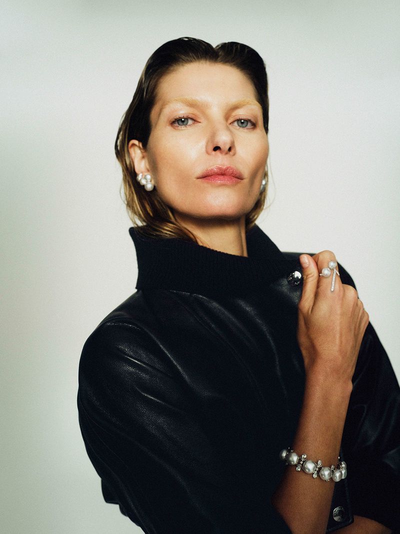 Portrait of a stylish adult woman with wet hair, wearing a black jacket and pearl jewelry against a neutral backdrop in a studio setting.