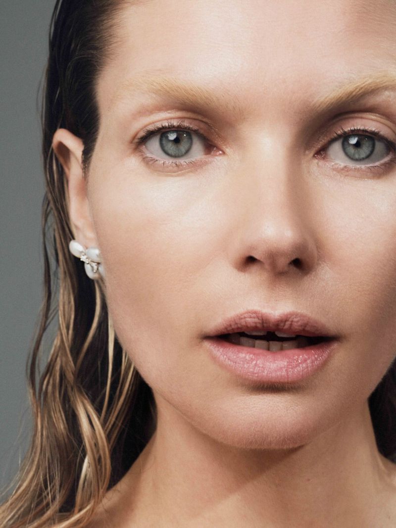Close-up portrait of a woman with wet blonde hair, pearl earrings, and light blue eyes against a solid grey background.