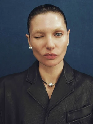 Model Jessica Hart winking while wearing a black leather jacket and pearl necklace against a dark blue background.