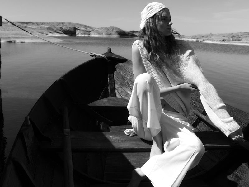 Monochrome fashion shot of a woman in white knitwear and a headscarf seated barefoot inside a wooden boat on calm water.