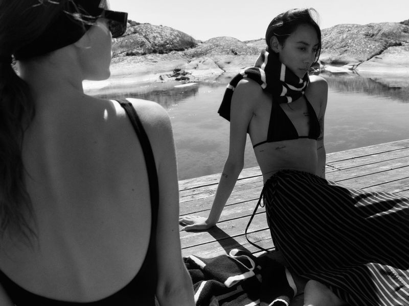 Two women resting on a wooden dock by rocky coastal waters in black swimwear and striped pieces in black and white.