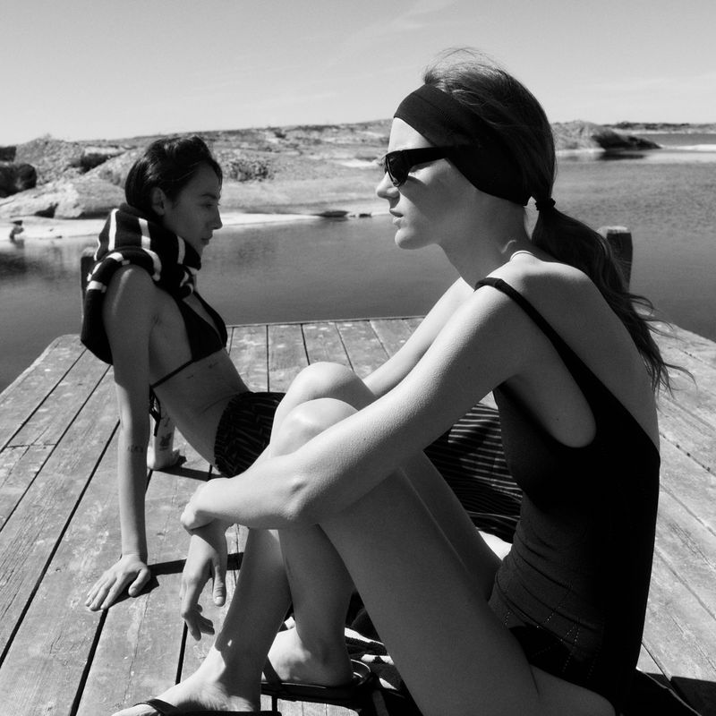 Two women in black swimwear and sunglasses relaxing on a wooden dock overlooking coastal waters in black and white.