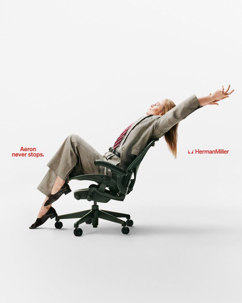 Woman in a grey suit reclining back in an ergonomic Herman Miller Aeron office chair against a white seamless background.