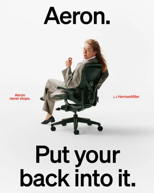 Model in an oversized grey suit sitting backwards on a dark Herman Miller Aeron chair against a white background.