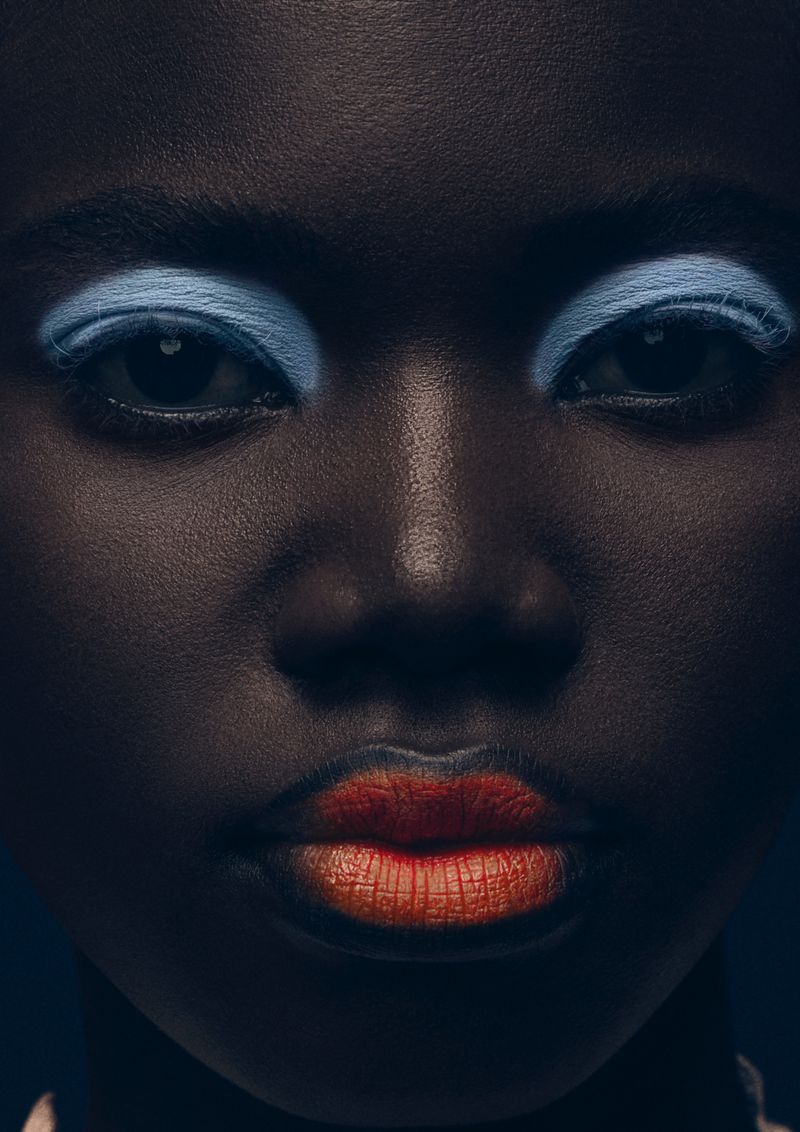 Close-up portrait of a Black woman with blue eyeshadow and orange lipstick, showcasing beauty and dramatic makeup artistry in studio lighting.