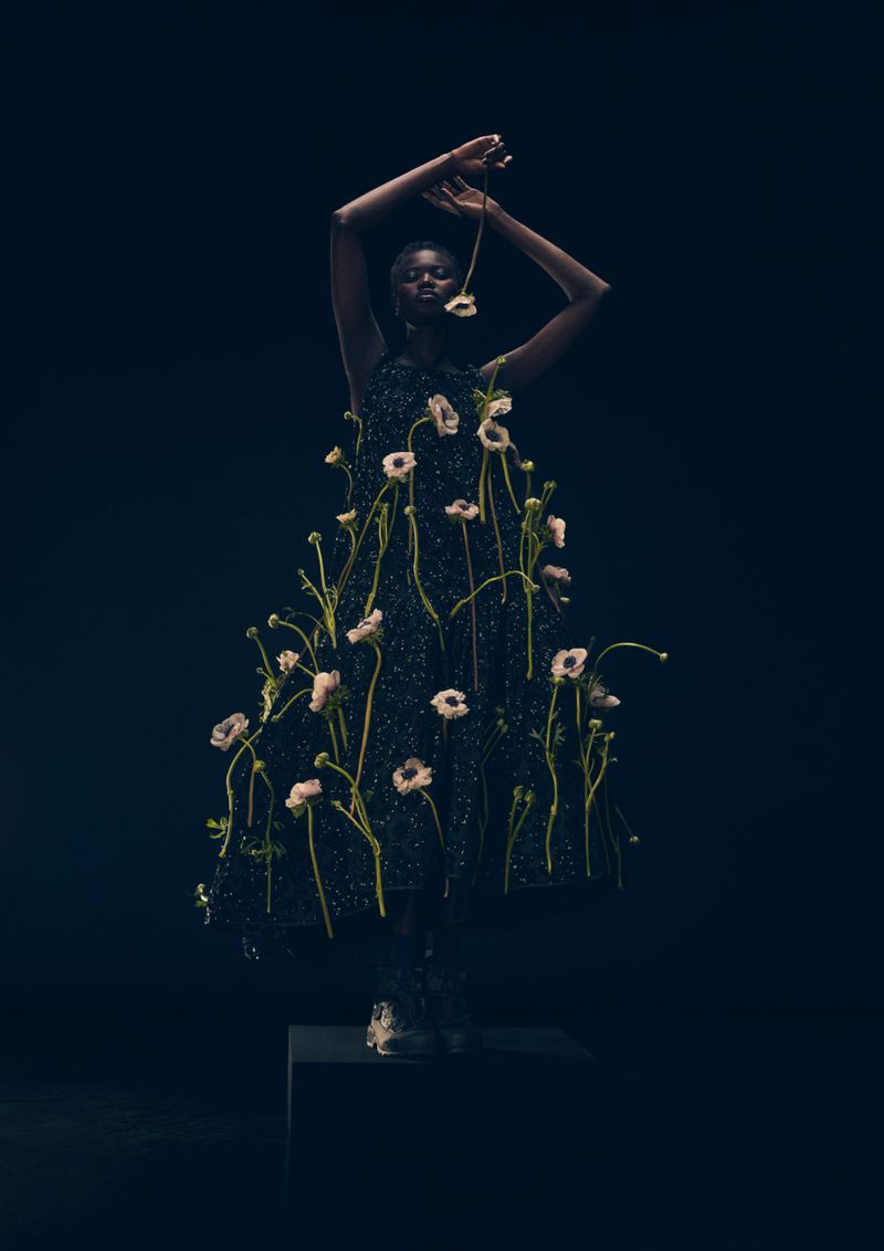 An African woman wears a dress adorned with fresh flowers, standing on a black cube against a dark background in a fashion portrait.