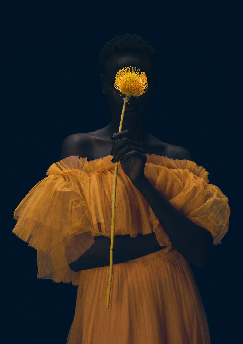 Woman with dark skin and a yellow dress covering her face with a yellow flower, set against a dark background in a portrait-style image.