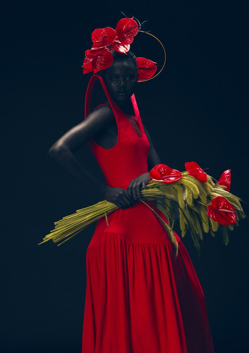 An adult female with black body paint poses in a red dress holding a bouquet of red anthurium flowers against a black backdrop.