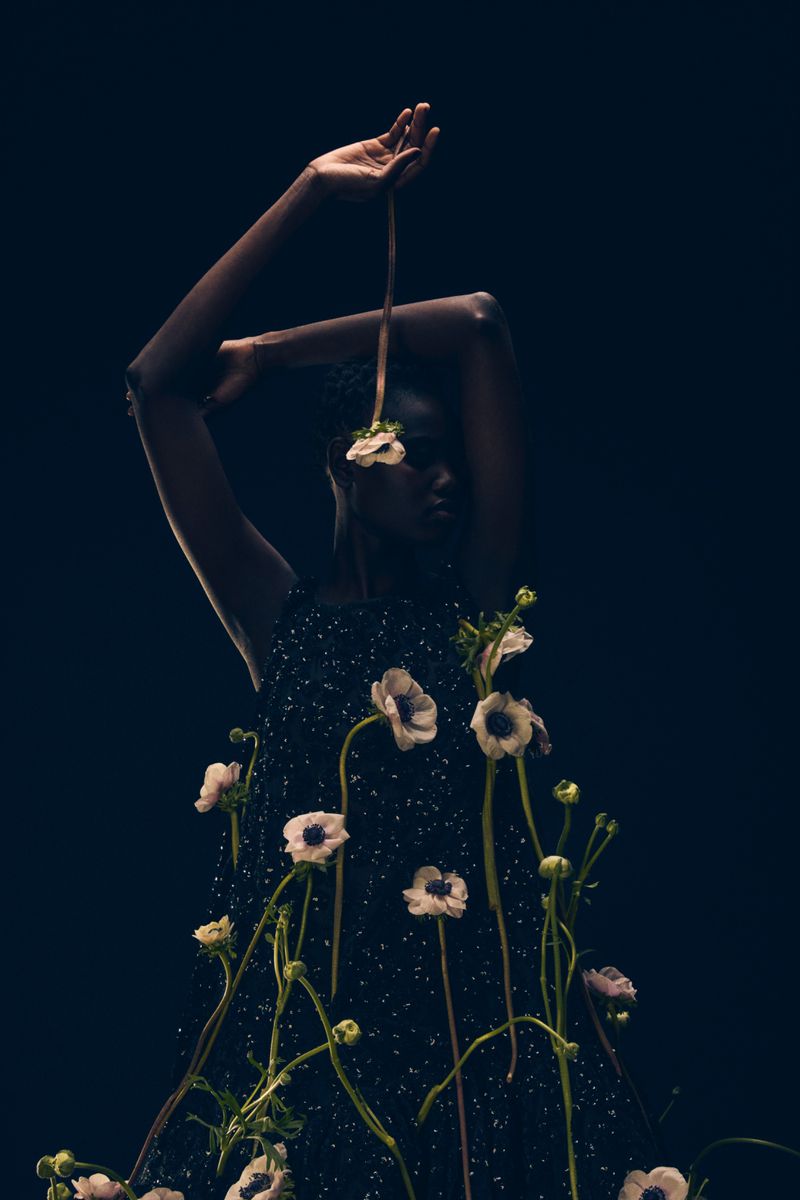 Fashion shot featuring a dark-skinned model in a textured black dress adorned with white anemone flowers in a dark indoor setting.