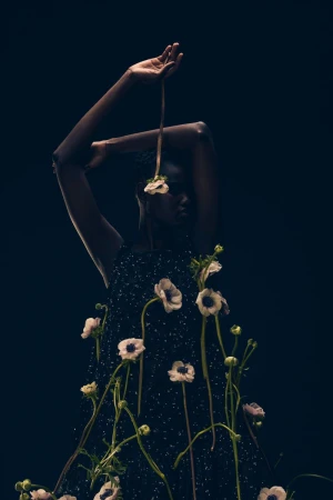 Fashion shot featuring a dark-skinned model in a textured black dress adorned with white anemone flowers in a dark indoor setting.