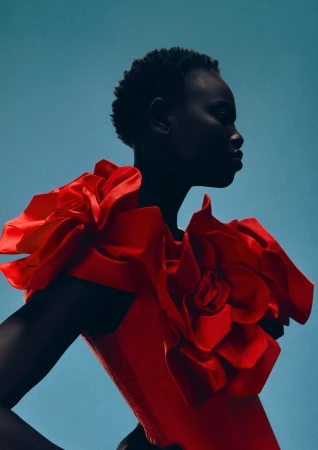 Profile shot of an African-descent woman wearing a red dress with large floral shoulder adornments, posing against a light blue backdrop.