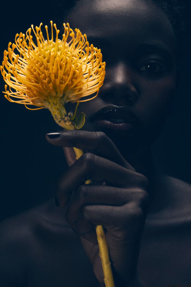 Portrait of a woman with dark skin holding a yellow pincushion protea flower, set against a dark, moody backdrop in a beauty fashion shot.