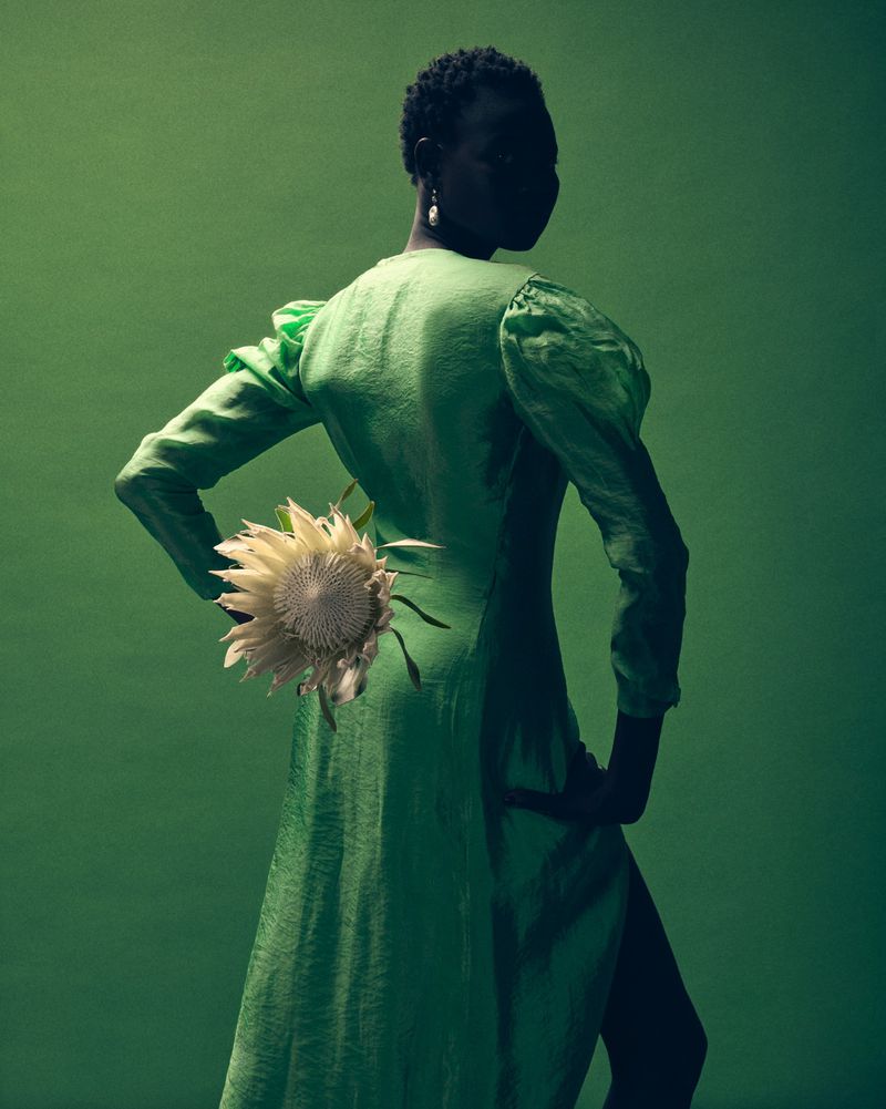 Elegant Black female model in green dress, holding a flower against a green backdrop in a studio setting, fashion editorial style.