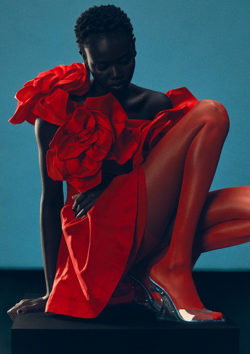 An African-American female model poses in a red dress with flower accents and clear heels against a blue backdrop in a studio setting.